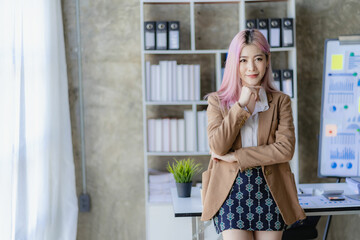 Attractive pink-haired Asian female employee standing in front of a desk Using a touch tablet computer, smiling, emailing business information to customers at a desk in the company's office.