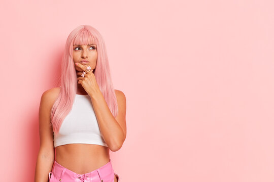 Young woman poses in the middle of pink wall of studio, stands with one hand on her chin, looks up thoughtfully waiting for good news, pause to think concept, copy space, high quality photo