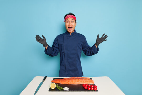Portrait photo of happy guy in cooking jacket and gloves at table against blue wall, hands with open palms, fish fillet with tomatoes and lemon in front of him, tasty food concept, copy space, high