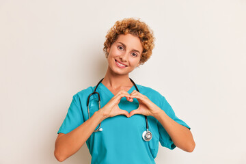 Portrait shot of pretty young woman with short curly hair wearing blue coat stethoscope on her neck, looking at camera with smile and showing heart sign with palms, health care concept, copy space