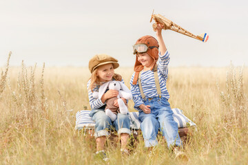 Boy and a girl are sitting on a suitcase in the grass under the open sky. Cheerful and happy children play in the field and imagine themselves to be pilots on a sunny summer day. Children dream of fly