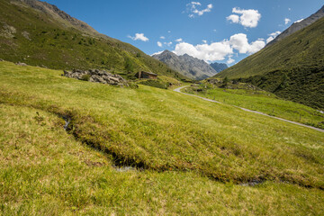 Naklejka premium View of an alpine landscape with a small river in the foreground, and a wooden bard with surrounding mountains in the background during summer. Umhausen, Tyrol, Tirol.