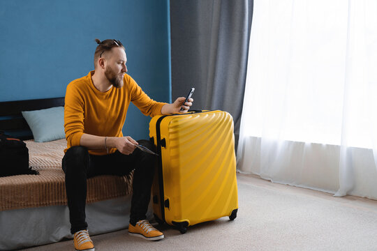 Young traveler in yellow sweater relaxing and using smartphone in hotel room after check in. Handsome man vacation travel blogger sitting on bed, holding phone for vacation, ready for trip. - Powered by Adobe