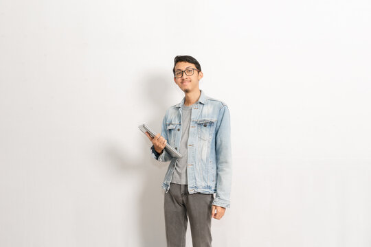 A guy looking confident wears a denim jacket with a laptop on his hand. Indonesian or southeast asian isolated model in front of white background.