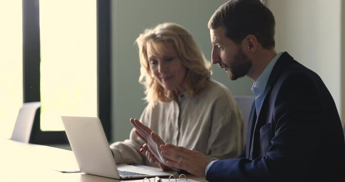 Serious confident business project leader explaining new employee work tasks, using laptop, pointing at display. Middle aged intern listening to speaking, mentor at workplace