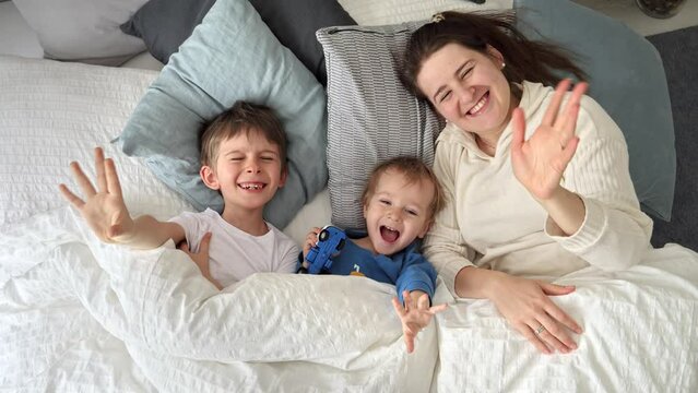 Young happy mother lying in bed with her two sons and waving in camera. Concept of family happiness, relaxing at home, having fun in bed, parent and cheerful kids