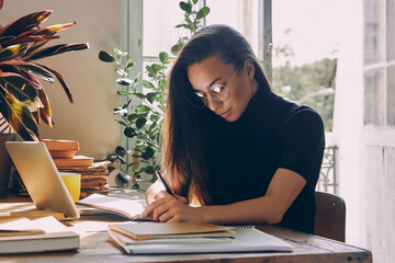 Confident young woman sitting at the desk and making notes while working from home