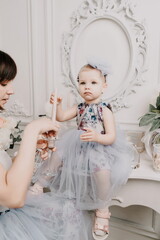 Fototapeta na wymiar Baby girl elegant dress. A one-year-old girl in a puffy dress and a cute bow poses against the backdrop of a bright room with a dressing table and flowers.