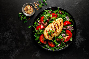 Chicken breast fillet grilled and fresh vegetable green salad with arugula, tomatoes and olives on black background, healthy food, mediterranean diet, top view - obrazy, fototapety, plakaty