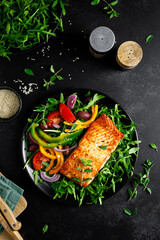 Salmon fillet grilled and fresh vegetable green salad of arugula with tomatoes, olives and bell pepper on black background, healthy food, mediterranean diet, top view