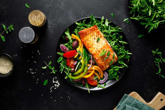 Salmon fillet grilled and fresh vegetable green salad of arugula with tomatoes, olives and bell pepper on black background, healthy food, mediterranean diet, top view