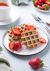 Homemade soft waffles with fresh strawberries and powdered sugar in a plate on a light background with a cup of tea. Traditional Belgian waffles.