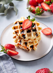 Homemade soft waffles with fresh strawberries and chocolate topping in a stack in a plate on a light background close up. Traditional Belgian waffles. Healthy breakfast.