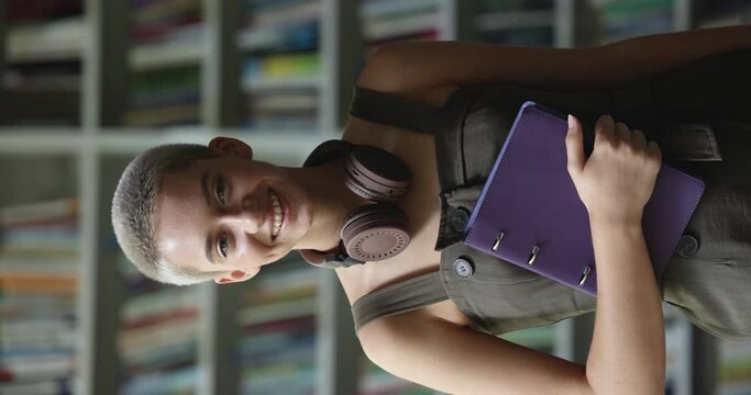 Pretty teen girl with short blond hair smile looks at camera posing in library on bookshelves background, vertical shot. Portrait of high school student, excellent studies, higher education, courses