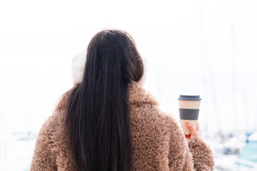 Young woman wearing winter muffs  and holding take away coffee at outdoors in back position