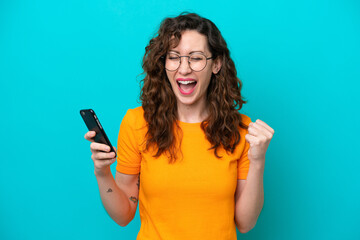 Young caucasian woman isolated on blue background using mobile phone and doing victory gesture