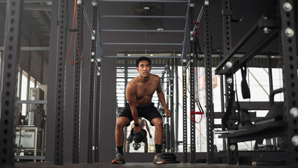 Strong asian man doing exercise with kettlebell at crossfit gym. Athlete male wearing sportswear workout on grey gym background with weight and dumbbell equipment. Healthy lifestyle.