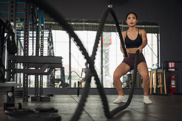 Obraz na płótnie Canvas Strong asian woman doing exercise with battle rope at crossfit gym. Athlete female wearing sportswear workout on grey gym background with weight and dumbbell equipment. Healthy lifestyle.
