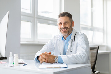 Fototapeta na wymiar Portrait of happy cheerful man in white workwear doctor posing at clinic, sitting at workdesk and smiling at camera