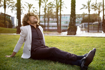 Obraz premium Relaxed african american middle aged businessman resting and sitting on grass in park, feeling free, escaping from work