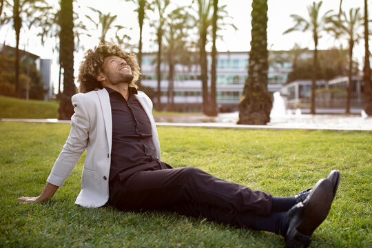 Relaxed african american middle aged businessman resting and sitting on grass in park, feeling free, escaping from work