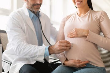 Obraz premium Healthcare, prenatal care. Male doctor examining pregnant woman's belly using stethoscope, listening baby's heartbeat