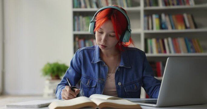 Assiduous student girl with dyed bright red hair listens audio course through headphones, improve foreign language knowledge, make notes studying in library. Igeneration use modern tech for education