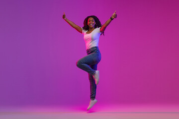 Fototapeta na wymiar Happy Black Lady Jumping Up In Neon Light And Showing Thumbs Up