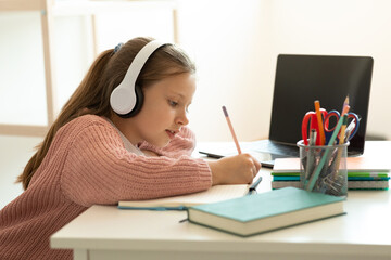 Busy european teenager girl in wireless headphones doing homework and listening lesson