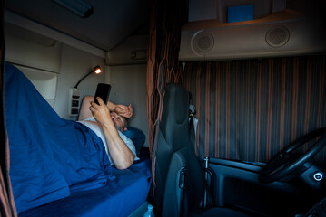A trucker in the cab,lying in the bed of the truck,scrolling on his phone during his off-time,with...