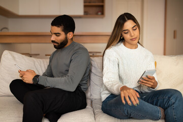 Sad offended millennial arab husband ignoring caucasian wife, look at smartphone after quarrel in living room