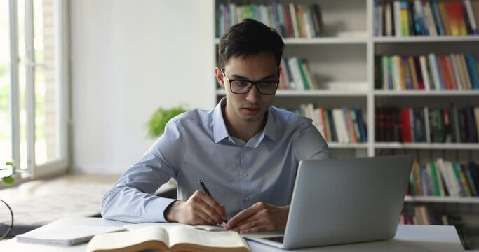 Indian teen student guy in glasses studying in university library, engrossed in creative task making, prepare for admission, doing coursework using laptop. Education, modern technology, intelligence