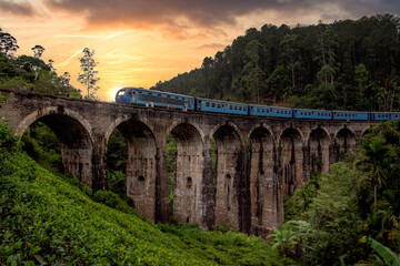 Fototapeta na wymiar A blue train crossed an old stone viaduct with the sunset in the background, surrounded by tea fields and mountains.