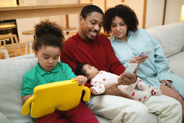 Happy loving multicultural family sitting on couch, using gadgets
