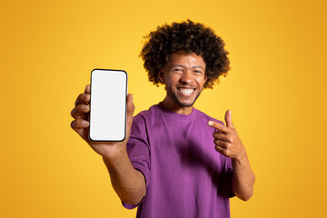 Obraz na płótnie Canvas Smiling adult african american curly guy in purple t-shirt pointing finger at smartphone with empty screen