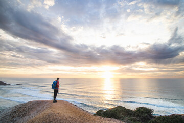 Traveler stands on the edge of a cliff near the town of Odeceixe, Algarve region, Portugal watching the sun setting on the clear blue ocean. The play of colors in the sky