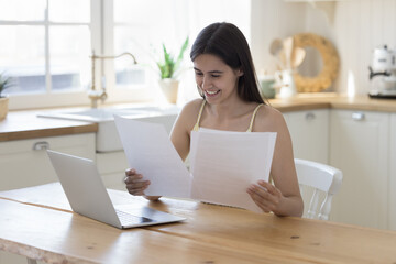 Happy young Latin woman reading paper document at home, doing paperwork at table with laptop...