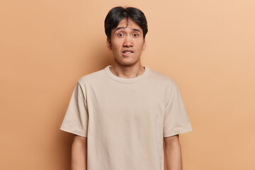 Waist up shot of worried Japanese man bites lips looks anxious and surprisingly at camera doesnt know way out from difficult situation dressed in casual t shirt isolated over brown background
