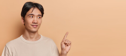 Horizontal shot of handsome brunet Asian man with short hair points index finger on blank space shows promo banner dressed in casual t shirt isolated on brown background. Place your advertisement here