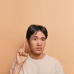 Vertical shot of serious Japanese man with dark hair points index finger above advertises something asks to look at ceiling dressed in casual t shirt isolated over brown wall. Advertisement concept.