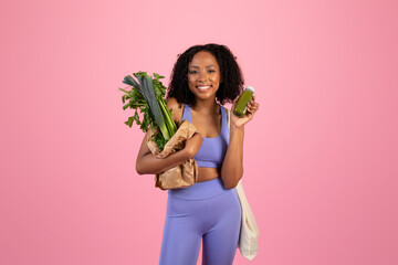 Smiling millennial black female in sportswear show paper bag with green vegetables and smoothie bottle