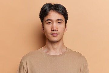 Portrait of serious dark haired adult Asian man with neutral facial expression focused at camera dressed in casual jumper isolated over brown background. Handsome Japanese guy poses in studio - 595545902