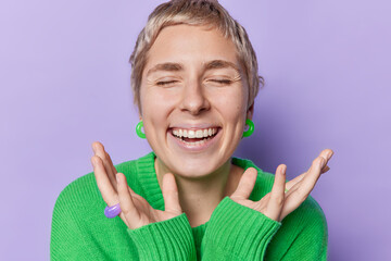 Portrait of short haired woman keeps eyes closed smiles broadly shows white teeth keeps hands near face wears green warm jumper and earrings isolated over purple background. Happy emotions concept