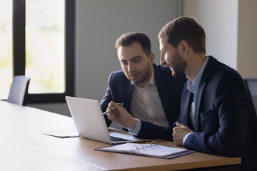 Two positive business colleagues men testing new professional application, watching online content, pointing at laptop display. Manager, developer showing software product to client, customer