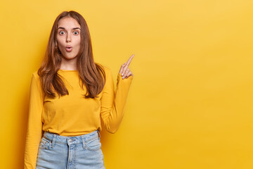 Studio shot of dark haired woman stares speechless at camera has opened mouth points finger at copy...