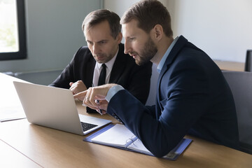 Two focused businessmen staring at laptop display at office work table, discussing online application, finding problem, watching software product presentation on Internet, showing, pointing at screen