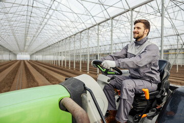 A happy agronomist is cultivating the land in hothouse while driving tractor.