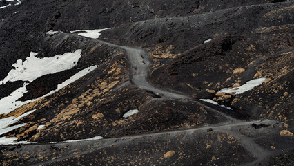 Snow melts on Etna volcano in spring. Panorama of uneven ground full of lava earth and craters. Small shrubs and large furrows, mediterranean climate. Colorful mountain trails on southern slope.