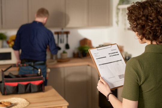 Rear view of young woman examining repair service contract while standing in the kitchen with repairman in background