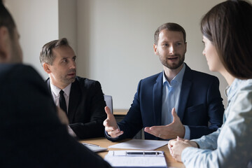 Positive confident ambitious young business leader man in formal suit presenting project to team, partners, investors on corporate meeting, speaking with hand gestures